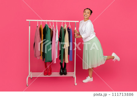 Smiling Asian lady standing beside garment rack filled with trendy clothes Smiling Asian lady standing beside garment rack filled with trendy clothes 111713886