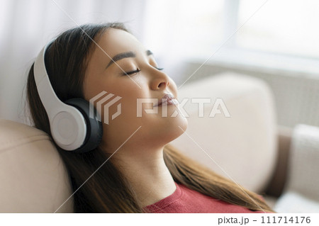 Calm Smiling Young Asian Woman In Wireless Headphones Listening Music At Home 111714176