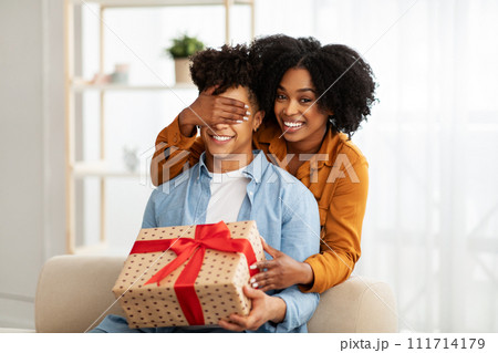 A woman surprises a man by covering his eyes from behind as they both smile A woman surprises a man by covering his eyes from behind as they both smile 111714179