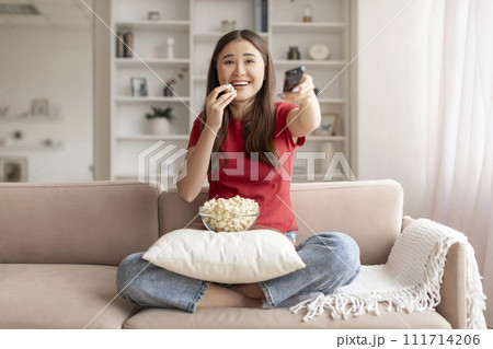 Cheerful young asian woman eating popcorn and holding remote control Cheerful young asian woman eating popcorn and holding remote control 111714206