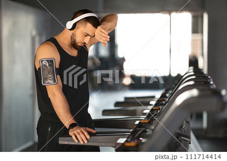 Exhausted young athlete wipes sweat from his forehead after treadmill session 111714414