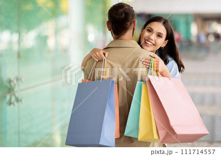 Cheerful millennial caucasian lady hug man shopaholic with many packages, enjoy shopping together, in mall Cheerful millennial caucasian lady hug man shopaholic with many packages, enjoy shopping together, in mall 111714557