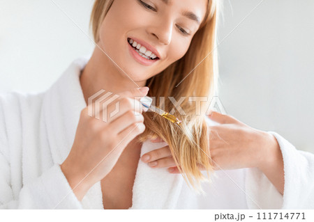 Blonde lady in bathrobe applies nourishing serum to hair indoor 111714771