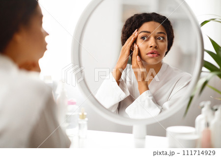 Sad african american woman looking in mirror at wrinkles around her eyes 111714929