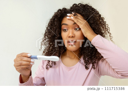 Surprised young black woman holding pregnancy test and looking with disbelief Surprised young black woman holding pregnancy test and looking with disbelief 111715386