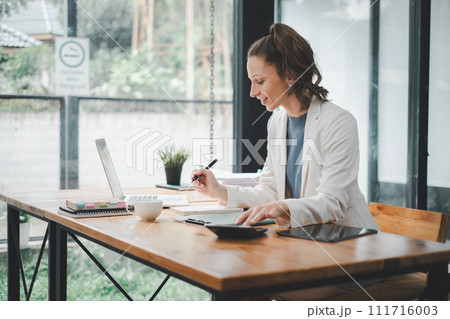 Business woman concept, A dedicated professional is immersed in work, writing in a notebook with a laptop and tablet on her well-organized office desk. 111716003