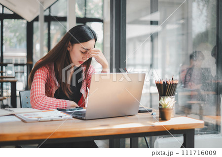 Business analytics concept, A young woman appears stressed as she works intently on her laptop at a wooden desk, surrounded by office supplies. Business analytics concept, A young woman appears stressed as she works intently on her laptop at a wooden desk, surrounded by office supplies. 111716009