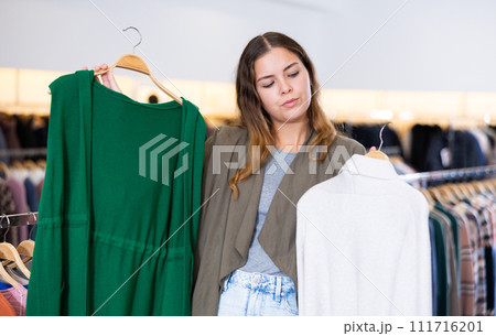 Young female choosing between green cardigan without sleeves and white sweater in a clothing store Young female choosing between green cardigan without sleeves and white sweater in a clothing store 111716201