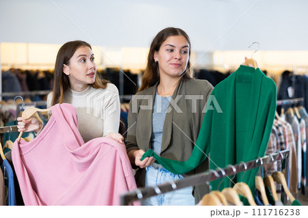 Positive young women testing fabrics of green and pink cardigans while shopping in a showroom Positive young women testing fabrics of green and pink cardigans while shopping in a showroom 111716238