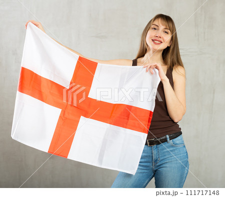 Joyful positive patriotic young woman holding state flag of England against gray wall background indoors 111717148