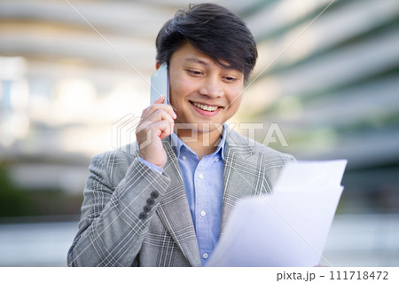 Asian businessman speaking by cellphone while holding documents on street Asian businessman speaking by cellphone while holding documents on street 111718472