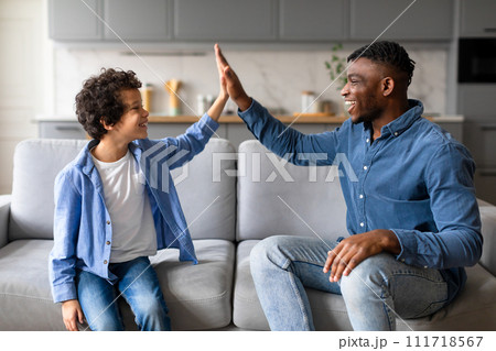Joyful black father and son high-fiving, celebrating happy moment on the couch 111718567