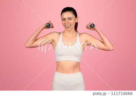 A cheerful woman flexes her muscles with a confident smile, holding small dumbbells A cheerful woman flexes her muscles with a confident smile, holding small dumbbells 111718666
