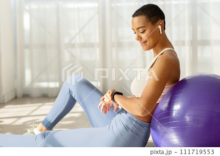 Positive young slim latin woman in sportswear, wireless headphones, check tracker pulse 111719553