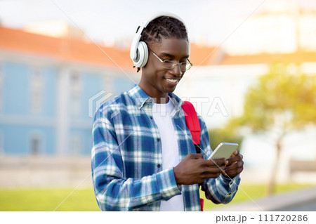 African student guy on cellphone listening audiobooks with earphones outdoors African student guy on cellphone listening audiobooks with earphones outdoors 111720286
