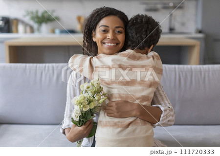 Smiling black mother receiving hug from son and holding bouquet of flowers Smiling black mother receiving hug from son and holding bouquet of flowers 111720333