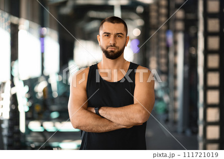 Confident muscular man in sleeveless top standing with crossed arms at gym 111721019
