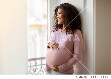 Black pregnant woman gently cradling belly while standing by window at home 111724149