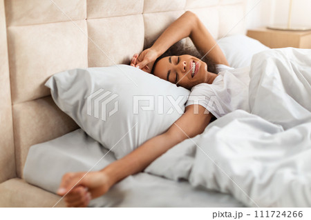 Positive black woman in white t-shirt stretching arms in bedroom Positive black woman in white t-shirt stretching arms in bedroom 111724266