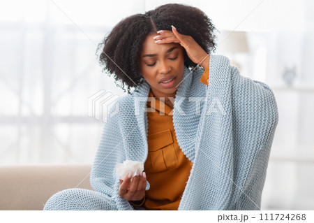 Distressed young woman with curly hair sitting on a couch, wrapped in a blue blanket Distressed young woman with curly hair sitting on a couch, wrapped in a blue blanket 111724268