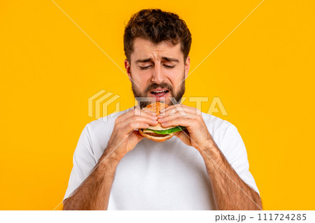 Unhappy bearded young man eats burger over yellow backdrop Unhappy bearded young man eats burger over yellow backdrop 111724285