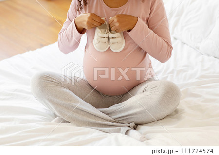 Pregnant lady sitting cross-legged on bed and holding pair of baby shoes 111724574