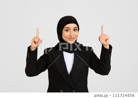 Smiling young businesswoman in hijab pointing upwards Smiling young businesswoman in hijab pointing upwards 111724856