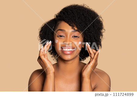 Happy black woman applying facial cream on cheeks and smiling Happy black woman applying facial cream on cheeks and smiling 111724899
