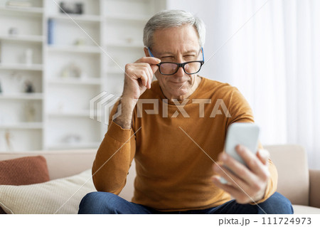 Elderly man adjusting his eyeglasses while using smartphone at home Elderly man adjusting his eyeglasses while using smartphone at home 111724973