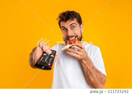 man enjoys slice of pizza while pointing remote control, studio man enjoys slice of pizza while pointing remote control, studio 111725242