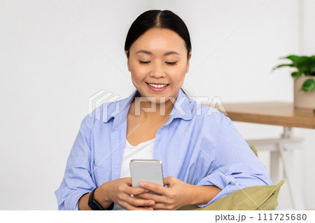 Positive korean lady enjoying weekend at home, using smartphone 111725680
