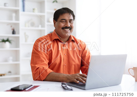 Cheerful mature indian businessman working on laptop at office Cheerful mature indian businessman working on laptop at office 111725750