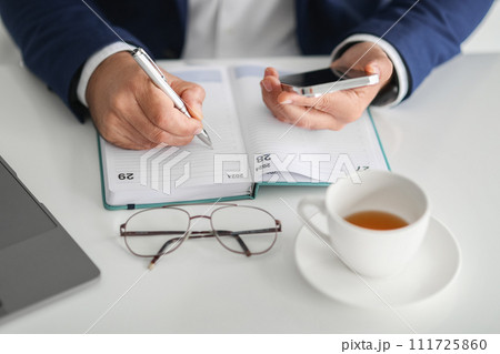 A businessman's hands are shown, one writing in an open planner and the other A businessman's hands are shown, one writing in an open planner and the other 111725860