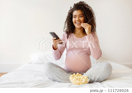 Pregnant Female Resting On Bed At Home, Watching Tv And Eating Popcorn 111726026