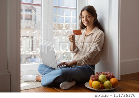 Smiling young woman sitting on floor with laptop and holding credit card Smiling young woman sitting on floor with laptop and holding credit card 111726376