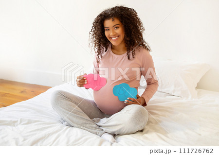 Cheerful expecting black mother sitting on bed with pink and blue signs Cheerful expecting black mother sitting on bed with pink and blue signs 111726762