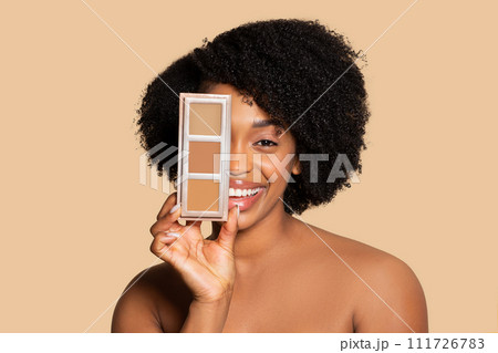 Happy young black woman holding concealer palette near face Happy young black woman holding concealer palette near face 111726783