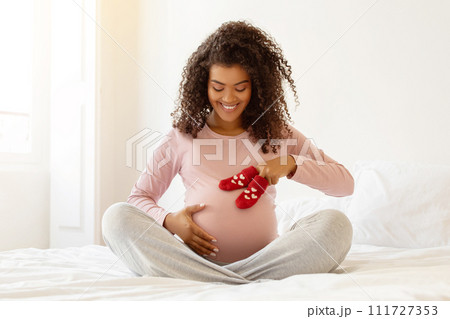 Black expectant mother sitting on bed and placing baby socks on belly Black expectant mother sitting on bed and placing baby socks on belly 111727353