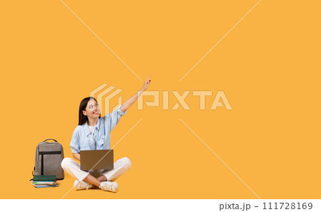 Student lady with laptop pointing upwards at free space sitting beside backpack Student lady with laptop pointing upwards at free space sitting beside backpack 111728169