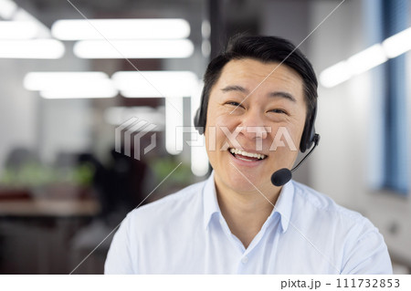 Webcam view, Asian support worker in shirt with headset smiling and looking at camera, businessman working inside office, consulting clients remotely, remote meeting with colleagues partners. 111732853