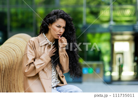 Worried woman coughing outdoors, feeling unwell. Health, sickness, public concern concepts in urban setting. 111735011