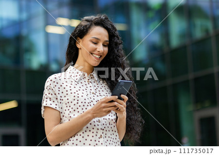 Young, attractive Hispanic businesswoman with curly hair checks her phone with a smile while taking a leisurely walk during her lunch break. Young, attractive Hispanic businesswoman with curly hair checks her phone with a smile while taking a leisurely walk during her lunch break. 111735017