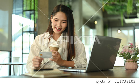 Happy young woman holding paper cup and looking at credit card while shopping online Happy young woman holding paper cup and looking at credit card while shopping online 111735103