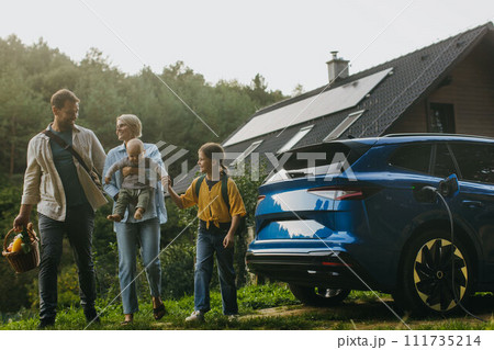 Family with electric car standing in front their house with solar panels on roof. Solar energy and sustainable lifestyle of young family. Concept of green energy and sustainable future for next 111735214