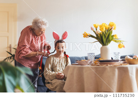 Grandmother braiding hair of granddaughter, putting ribbon ob braid. Prepairing for easter lunch. Happy easter. Grandmother braiding hair of granddaughter, putting ribbon ob braid. Prepairing for easter lunch. Happy easter. 111735230