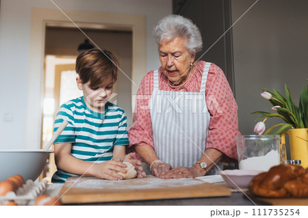 Grandmother with grandson preparing traditional easter meals, kneading dough for easter cross buns. Passing down family recipes, custom and stories. Grandmother with grandson preparing traditional easter meals, kneading dough for easter cross buns. Passing down family recipes, custom and stories. 111735254