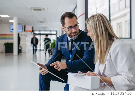 Pharmaceutical sales representative talking with doctor in clinic lobby. Ambitious male sales representative presenting new medication, drugs, showing data on tablet. 111735410