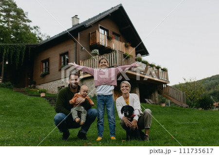 Family standing in front their house with solar panels on roof. Solar energy and sustainable lifestyle of young family. Concept of green energy and sustainable future for next generations. Family standing in front their house with solar panels on roof. Solar energy and sustainable lifestyle of young family. Concept of green energy and sustainable future for next generations. 111735617