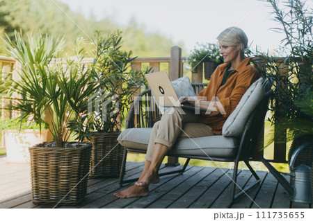 Woman working in the garden, with laptop on legs. Businesswoman working remotely from outdoor homeoffice, eating light lunch, sandwich. Concept of outdoor home office. Woman working in the garden, with laptop on legs. Businesswoman working remotely from outdoor homeoffice, eating light lunch, sandwich. Concept of outdoor home office. 111735655