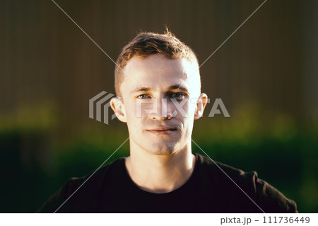 Male exterior portrait in evening lighting, close-up face of rugged white male in his 20s looking into camera against blurred rural landscape. 111736449